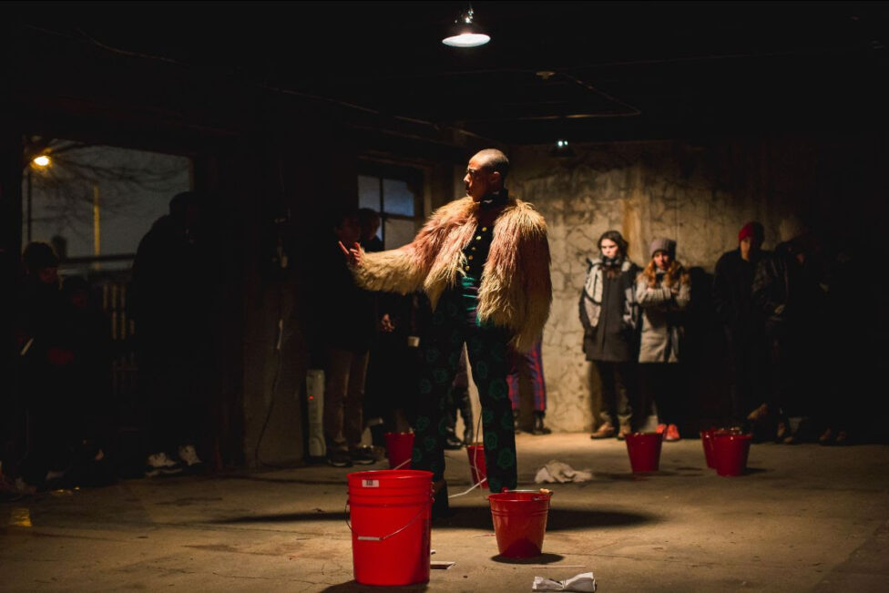 Photo of Fana Fraser performing. She is standing in a low lit room wearing a cream and light pink fur coat. She is standing with one arm slightly raised and one finger pointing upwards. There are red buckets placed around the room. Photo by David Gonsier. 