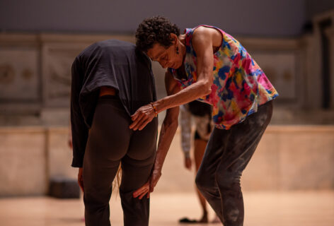 A photo of Barbara Mahler guiding a student during her class. The student faces away from the camera in a soft forward fold, letting their shoulders and head fall forwards. Barbara places a hand at the student’s sacrum and behind their thigh. Barbara wears a bright multicolored top with pink, blue and yellow patterns. Photo by Rachel Keane.
