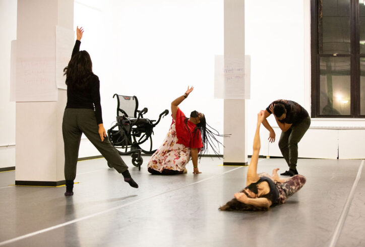 [ID: Four artists in a white studio with grey Marley floor. First artist on the left in dark clothing reaching up to the sky with one hand, standing leg bent while other leg is pointing and reaching to the side. Second artist, wearing black leggings, a light flower dress, red short sweater, and black mask is on their knees, one hand touching the ground, the other moving to reach towards the sky next to a wheelchair. Third artist, dressed in dark clothing and mask, is rounded over, touching their shoulder while gazing and reaching towards the ground. Final artist, dressed in flower leggings, black tank top, and mask, lays on the floor, with one leg in passé, foot flexed, reaching towards the sky with hair loose and splayed on the floor. Behind the artists, reads a sign on a white column 'What are you again?']