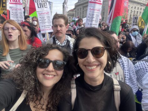 Photo of Alia (left) and Amany (right) at a protest for Palestine. Both are smiling brightly and wearing sunglasses and black tops. In the background protestors crowd around them holding signs and several wearing the keffiyehs. Photo by Alia Al-Sabi.