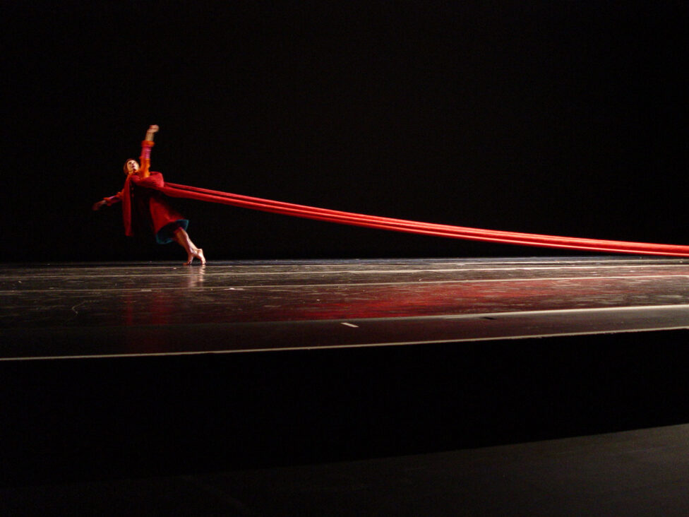 On a darkened stage, dancer Hristoula Harakas wears a red costume whose long train extends behind them. Photo courtesy of the artists. 
