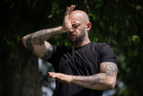 Christopher holds his right hand in between his eyebrows. The left one on transverse plane. He wears a black tee. His arms are heavily tattooed. Photo by Walter Wlodarczyk.