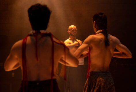 Graphic art with text over an image. Two dancers turn their backs and Christopher Unpezverde Núñez looks straight ahead. All three identify the solar plexus in their chests. An amber light bathes them. Photo by Maria Baranova.