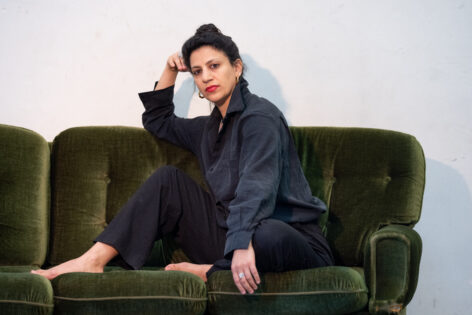 a portrait of Sahar Damoni. She has Black hair pulled back, and black T-shirt and Black Pants. She looks into the camera in neutral Expression with red lipstick on her lips. She sits on a green velvet couch. Her head rests on her right hand propped up on the couch. Her left hand rests on her left leg. Her legs are crossed and the background is white. Photo by Dirk Rose.