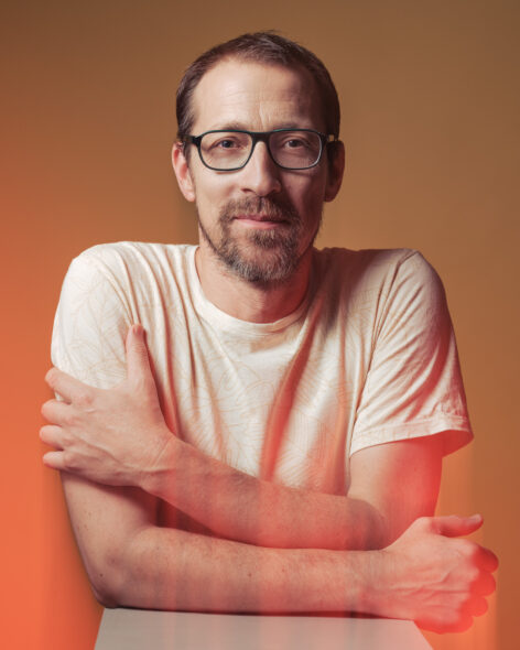 Portrait of Gergely Talló. He is wrapping his arms around his torso. He wears a white t-short and dark rectangle frame glasses. He looks straight into the camera. Photo by Domolky Daniel.