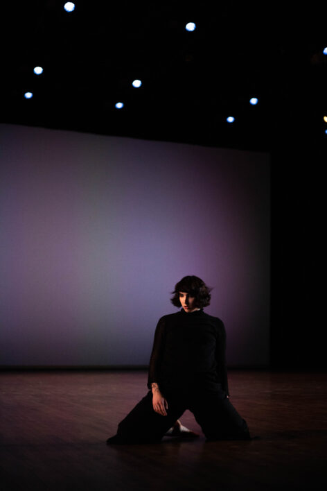 Brooke performs. She is kneeling on a wood floor, wearing a black turtleneck and black pants. Photo by Elisheva Pichanick.