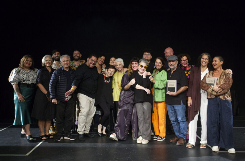 [A group of artists wearing modern clothing embrace each other as they stand on a stage with joyous expressions on their faces. Two people cradle copies of the book Habitar lo Imposible: Danza y experimentación en Puerto Rico in their hands]. Artists and collaborators at the book presentation in March 2023, Teatro Julia de Burgos, Universidad de Puerto Rico. Photo by Thais Llorca