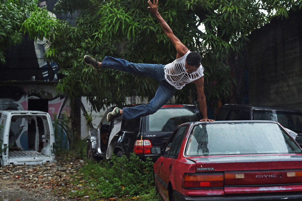 [Under a lush green tree sit a group of abandoned automobiles. Javier Cardona in a white tank top with cut-outs and denim jeans presses their left hand into a red car as they extend their limbs in the air]. Javier Cardona Otero, Hasta el cuello, 2016, Gandul community, San Juan, PR. Photo by Ricardo Alcaraz.