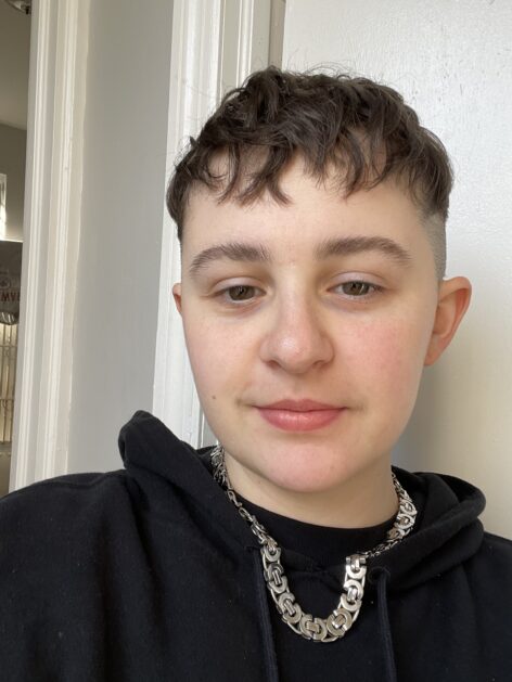 Portia, a white transmasculine person, appears wearing a black hoodie and silver chain necklace. Their hair is brown and cut with messy short bangs and shaved on the sides. They smirk at the camera. Photo courtesy of the artist.