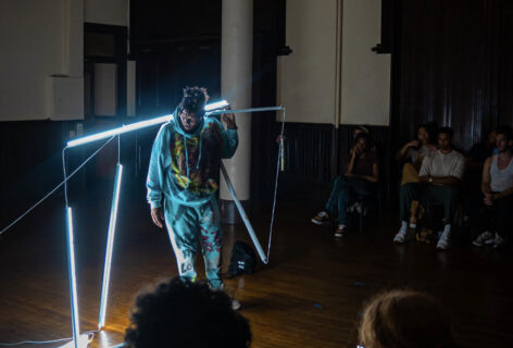 Photo of Malcolm x-Betts in a performance. They wear a teal hoodie and pants set that has been painted with yellow and orange shapes. They stand balancing several long LED lamps on their shoulders. several hang to the ground. In the foreground an audience is watching. Photo by Michelle Y. Thompson