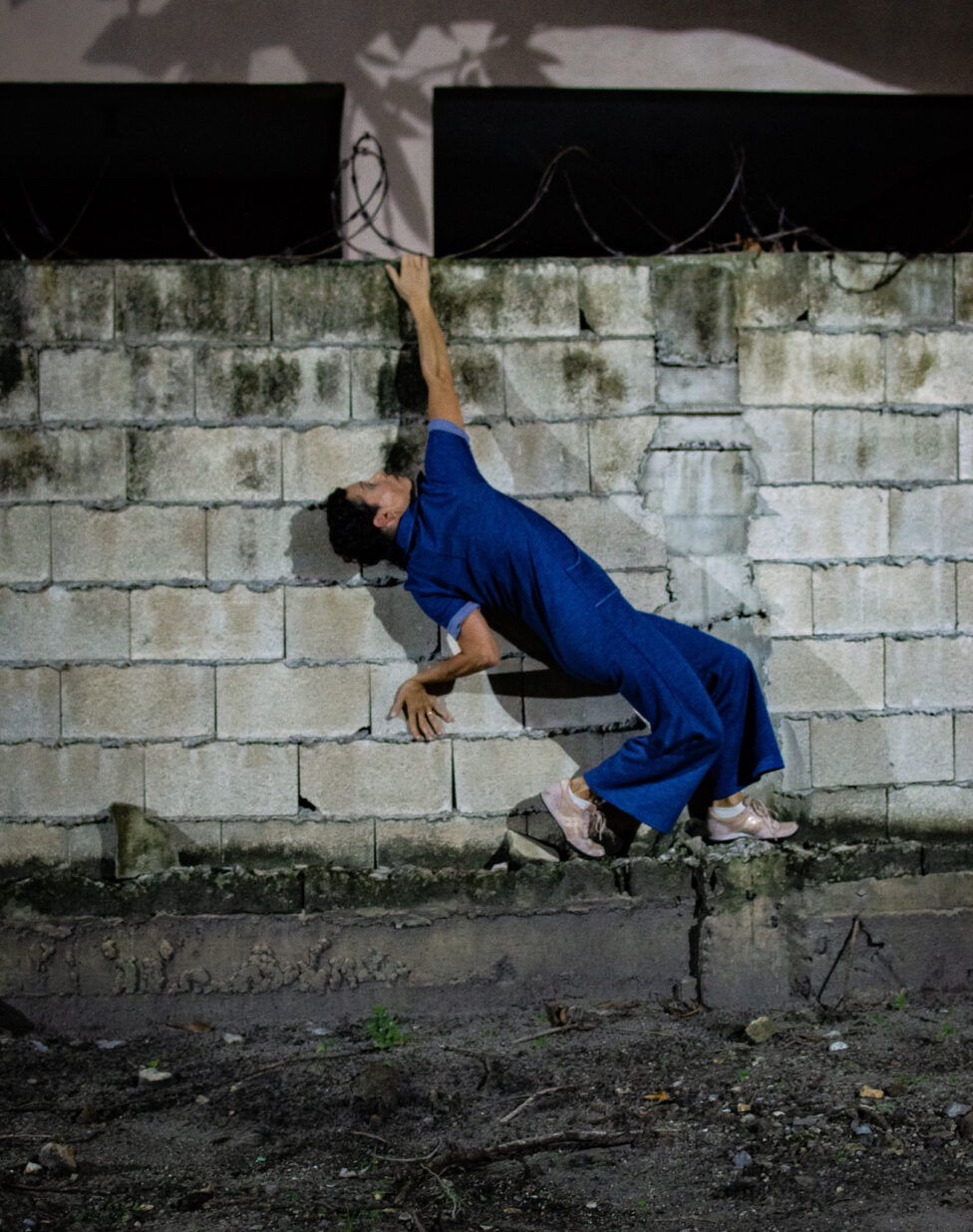[Karen Langevin wearing a blue jumpsuit and pink sneakers hoisting diagonally against a concrete brick wall]. Photo by Mariana Belaval 