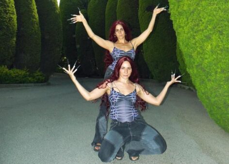 Noura and Salma pose for the camera with their arms extended out and elbows bent. Their palms are open and their fingers spread out like fans. They wear long transparent nails that reflect the sunlight. They are stacked in the photo, Noura sits while Salma stands behind. In the background there are large green bushes. Photo by Yazan El Zubi.