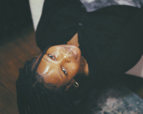 Photo of Marcelline lying down and looking up into the camera. She wears a black cardigan and her hair is in braids. Photo by Andina Clarkson.