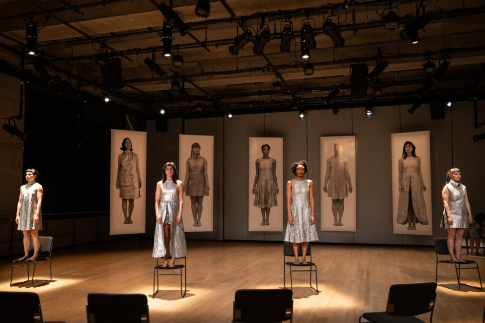 Dancers Pareena Lim, Molly Lieber, Bria Bacon, and Natalie Green wear iridescent gray dresses as they stand on evenly spaced black chairs under individual spotlights. Five full-length images of people wearing similar dresses hang on a wall in the background. Photo by Walter Wlodarczyk.