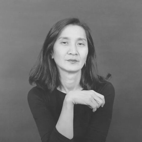 Black and white headshot of Donna Uchizono with dark, shoulder-length hair in a black top sitting with one elbow bent with her hand rising toward her chin.