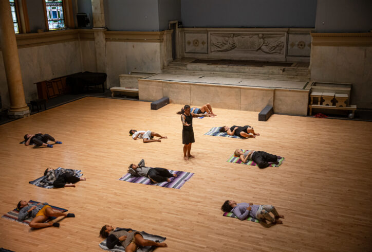 A photo of Fabio Tavares teaching a class at the Judson Church. He stands in the middle while students lay on woven blankets. Their knees are bent falling to one side while their faces turn to look away in opposition. Photo by Rachel Keane.