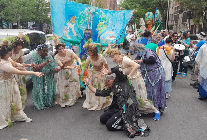 Several performers wearing robes and woven straw head dresses. They are on the street arms extending outwards reaching to a performer in black who is seemingly falling to the floor. Behind them a performer with their face painted in blue and wearing a teal long sleeves. They have a backdrop behind them of a painter earth. Photo courtesy of Ecological Cities.