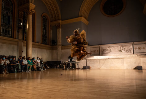 Photo of Maxi Hawkeye Canion. They are performing for Movement Research at the Judson Church. They wear a light brown costume made of many layers of tulle and a head piece with two long ears sticking straight up. They are in mid air, jumping, keens bent. In the background the audience looks on.