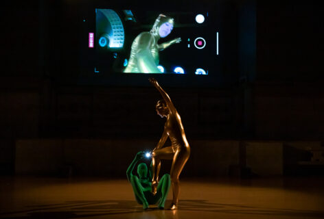 Photo of Katy Pyle performing for Movement Research at the Judson church. Two dancers in head to toe unitards. One dancer in green lays on the floor curving up and holding a cellphone with the flash on aimed at Katy. Katy wearing gold is standing balancing on one leg with the other leg bent and foot pointed. Their arm reaches up. On the projection above them we see the live feed being recorded of Katy from below. Photo by Rachel Keane.
