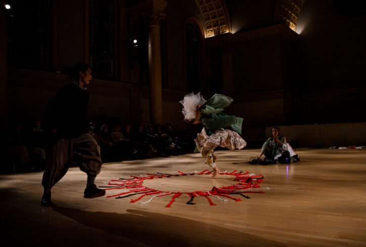 Photo from Ching-I Chang's performance for Movement Research at the Judson Church. In the center a dancer wearing a costume with many layers of white and green plastic ruffling all around them as they move. Two other performers look on, one stands and the other crouching. There is a circle of red fabric on the ground with red, white, and black strips shooting out of it.In the background the audience can be seen bearing witness. Photo by Rachel Keane.