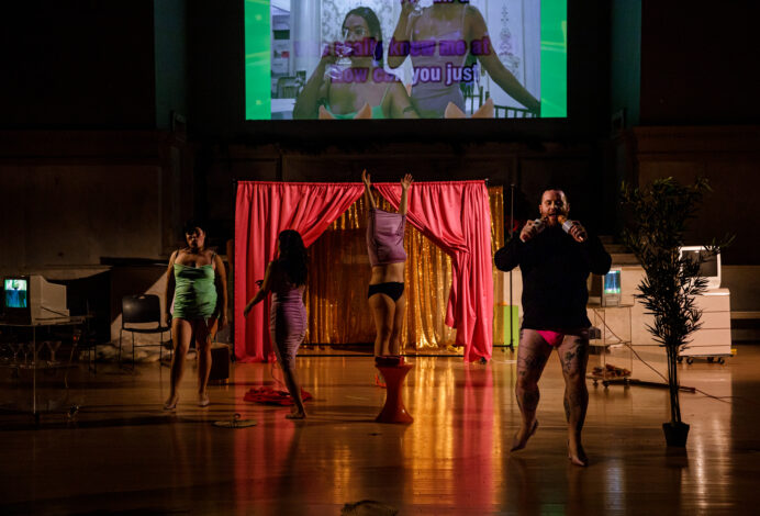 Photo from Kat Soltelo's performance for Movement Research at the Judson Church. A maximalist stage design with a projection, a pink theater curtain open to reveal a sparkly gold curtain. There are several TVs on carts around the stage as well as fake plants. The performers move around the space wearing tight fitting dresses or in various stages of undress. Photo by Rachel Keane.