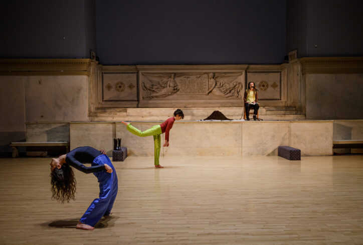 Photo of Leah Fournier, Amelia Heintzelman and Tess Michaelson performing for Movement Research at the Judson Church. Tess sits at the altar with headphones on. There is a pile of earth next to her. Lea wears bright green pants and balances on one leg. Torso moves forward while the other leg estends pack in counterbalance. Amelia stands in a deep back bend long hair reaching towards the floor. Photo by Rachel Keane.