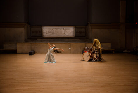 Stacy Lynn Smith and Alex Romania performing for Movement Research at the Judson Church. Stacy is kneeling wearing a light blue dress. Their right arm is bent hand to their chest while the other reaches out. Their head is tilted looking upwards and diagonally. Alex Romania sits at a drum set covered in long shiny gold plastic strips. Photo by Rachel Keane