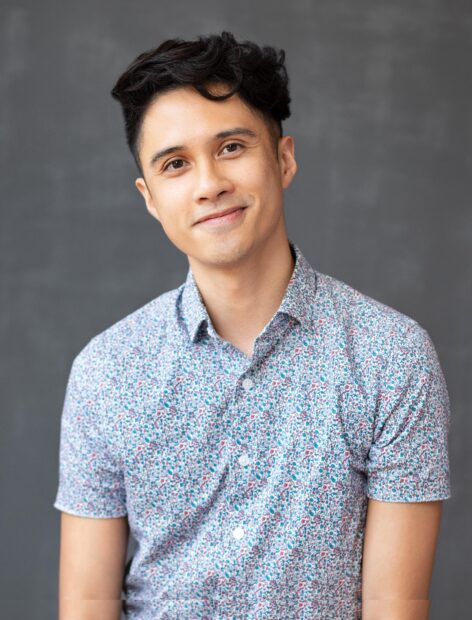 A portrait of a queer dancer with a medium complexion. He has short, wavy black hair, and his dark eyes are looking directly at the camera. He wears a collared, short-sleeved shirt with a colorful, intricate print. The backdrop is a plain, textured grey wall. Photo by by Tanya Rosen Jones.