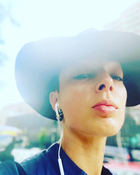Alicia is wearing a wide brimmed black hat looking down towards the camera with wired headphones in their ear. They are wearing a navy blue collared shirt with the city as their background.