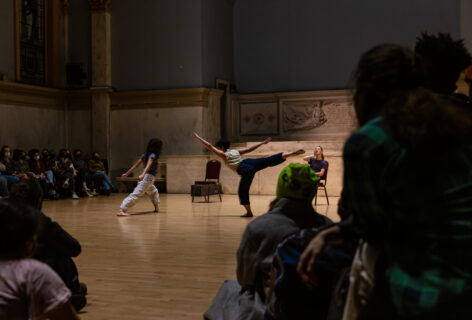 Photo of Julia Antinozzi performing for Movement Research at the Judson Church. In the foreground audience members looking forward towards the performers. One dancer sits at a chair. Another is in an arabesque position. The standing leg bends and the other reaches diagonally up. Another dancer in a deep lunge with their arms reaching straight back. Photo by Rachel Keane.