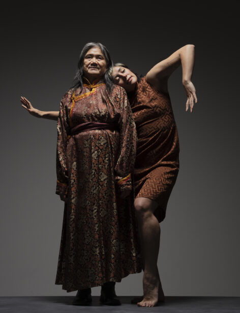 Melissa, a light-skinned and mixed race person leans their head on Joy's shoulder while extending one elbow diagonally upward and the other arm behind Joy. Joy, an older Chinese woman stands upright while looking out with a slight smile. They wear brown silk Chinese garments behind a gradient gray background.