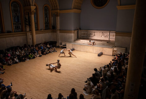 Company [REDACTED] performing for Movement Research at the Judson Church. The photo os taken from above. dancers are moving in the space. There are three duets and they all wear white. They push pull and climb on each other. Photo by Rachel Keane.