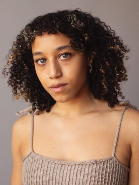 A curly-haired Brown-skinned woman with green eyes wears a beige tank top and stands in front of a grey wall. Photo by Avery Johnson.