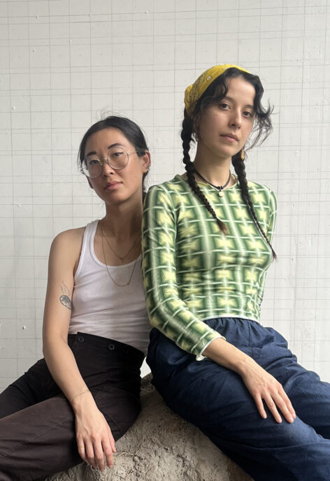 Portrait of Earthworks (Cristina Bartley Dominguez + Catherine Chen). both artists are seated on a rock. one artist wears a white tank top and brown slacks. They wear glasses and their hair is pulled back away from their face. The other wears a green and yellow square patterned top, navy blue pants and a yellow paisley print bandana. Their hair is in two braids. Both look into the camera with a neutral expression. Photo courtesy of the artists.