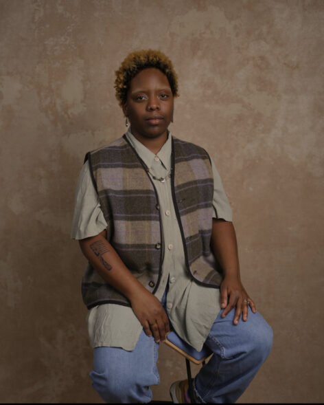 angel edwards, a brownskinned agender person sits in front of a neutral light pink background. They wear a brown tripped vest over a short sleeve gray button up top and blue jeans. Photo courtesy of the artist.