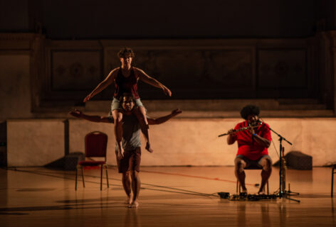 Photo of Julian Barnett's performance for Movement Research at the Judson Church. Two on dancer balances the other on their shoulders. They both hold their arms out for balance. On the right a musician plays a thin wind instrument. Photo by Rachel Keane.