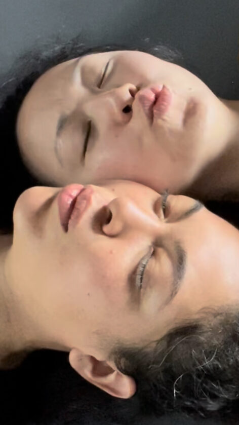 The heads of two women framed tightly. They are lying side by side horizontally, ear to ear, facing opposing directions. Both have their eyes closed. Both have dark hair. Their lips are puckered as if they are mid-song. Photo courtesy of the artists.