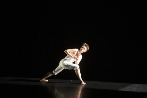 Cassie is wearing white leggings and a white tank top against complete darkness, illuminated by a single lane of white light. They are in a deep right lunge, torso tilted over their right thigh and head tilted outwards, with both arms crossed in front of them - left over right - and palms facing inward.