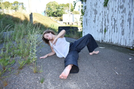 Dancer on gravel floor with green grass and grey distressed building in the background. Laying on right forearm, extending right leg out, left elbow bent up toward the sky, and left leg bent and reaching back, foot planted on the ground. Dancer is wearing a white flow tank-top and navy blue jeans.