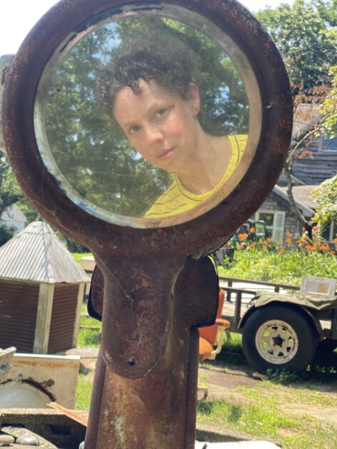 A round mirror with a rusted metal frame sits in a yard among lots of antique furniture and odd objects. In the mirror is Laurie's reflection: a woman with curly brown hair and a yellow shirt, half smiling and looking at the camera. Photo courtesy of the artist.