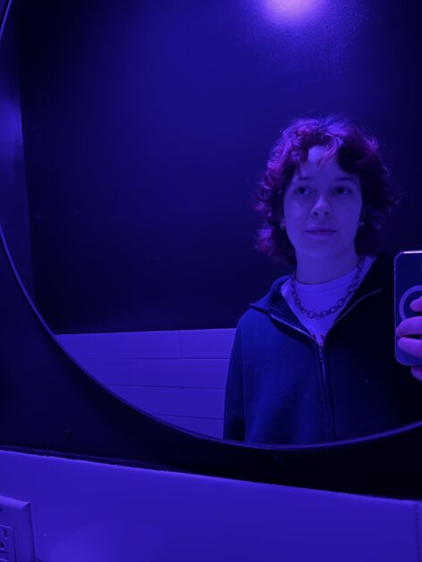 Artie is taking an off-center mirror selfie wearing a silver chain, white t-shirt, and navy blue zip up hoodie. The light in the bathroom casts a violet glow.