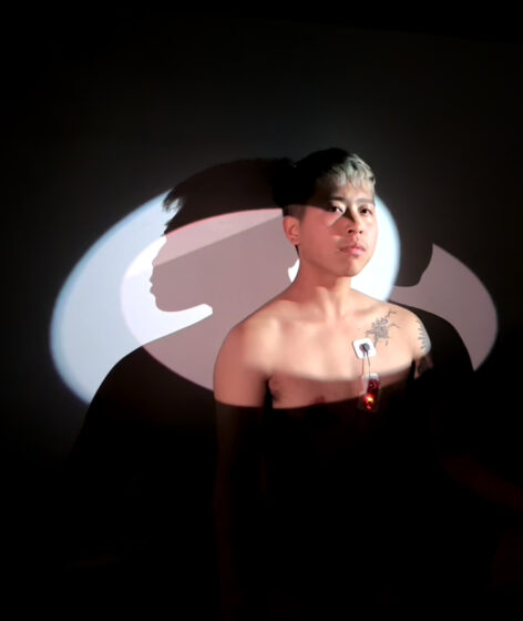 Portrait of Riven Ratanavanh in two overlapping beams of pale blue and pink light. The artist is shirtless, wearing a heart monitor attached to their chest, and looks into the camera with a defiant and pensive expression. Photo courtesy of the artist.