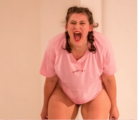 A photo of Emily Barasch she is facing the camera bent forward and wearing a pink t-shirt. She has pigtails and screams with her mouth wide open. Photo courtesy of the artist.