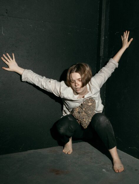 Color film photograph of Shannon Brooks crouching with arms and hands extend out and behind them. Their eyes are closed. They are barefoot. In their lap is a large, jagged, rock. Shannon is a white person with short brown hair with a horizontal blonde stripe across the middle of their head. Photo by Chris Postlewaite.