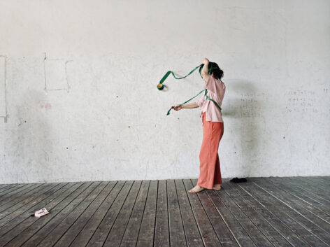 A photo of Zhangxinan standing on a wooden floor against a wall with writings on it. She interacts with a green ribbon-like tape that spirals around her body, one end attached to the wall. She is wearing a loose-fitting light-pink shirt and peach-colored trousers, with her face obscured by the raised arm. Photo by Yongxin Fu.