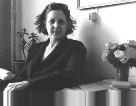 Photo of Nina Winthrop seated on a couch. The photo is in black and white. She wears a dark blazer and looks into the camera with a slight smile. She has short dark hair. Photo courtesy of the artist.