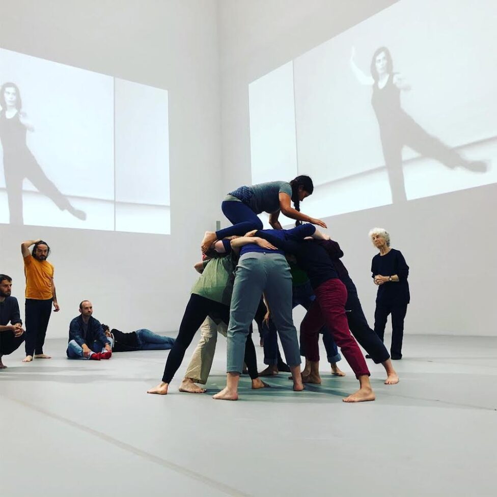 Martita (top) rehearsing “Huddle” with Simone Forti (right) and the cast of Dance Constructions at the Museum of Modern Art. Projected video of young Yvonne Rainer along the wall.
