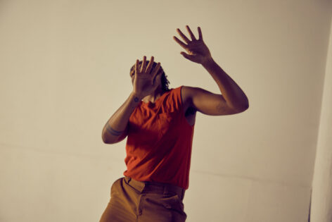 Jordan Demetrius Lloyd stands with his palms pressing outwards towards the camera. His hands obscures his face. He wears a burnt orange tank and brown pants. Photo by Whitney Browne.
