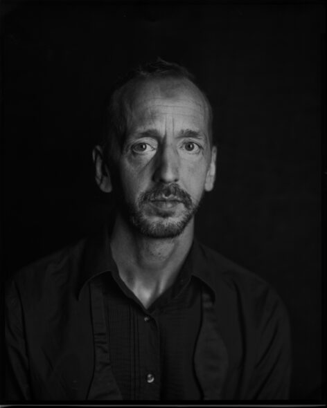 Black and white portrait of John Jasperse. He has a mustache and wears a dark colored top. He looks into the camera with a neutral face expression. Photo by Tei Blow.