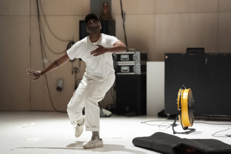 Jordan Demetrius Lloyd, dressed in all white looks up as his right hand extends to the back, with his left extended out in front of him. He stands on a white floor next to a yellow fan, and a thick black material folded up. Photo by Thomas Lenden.